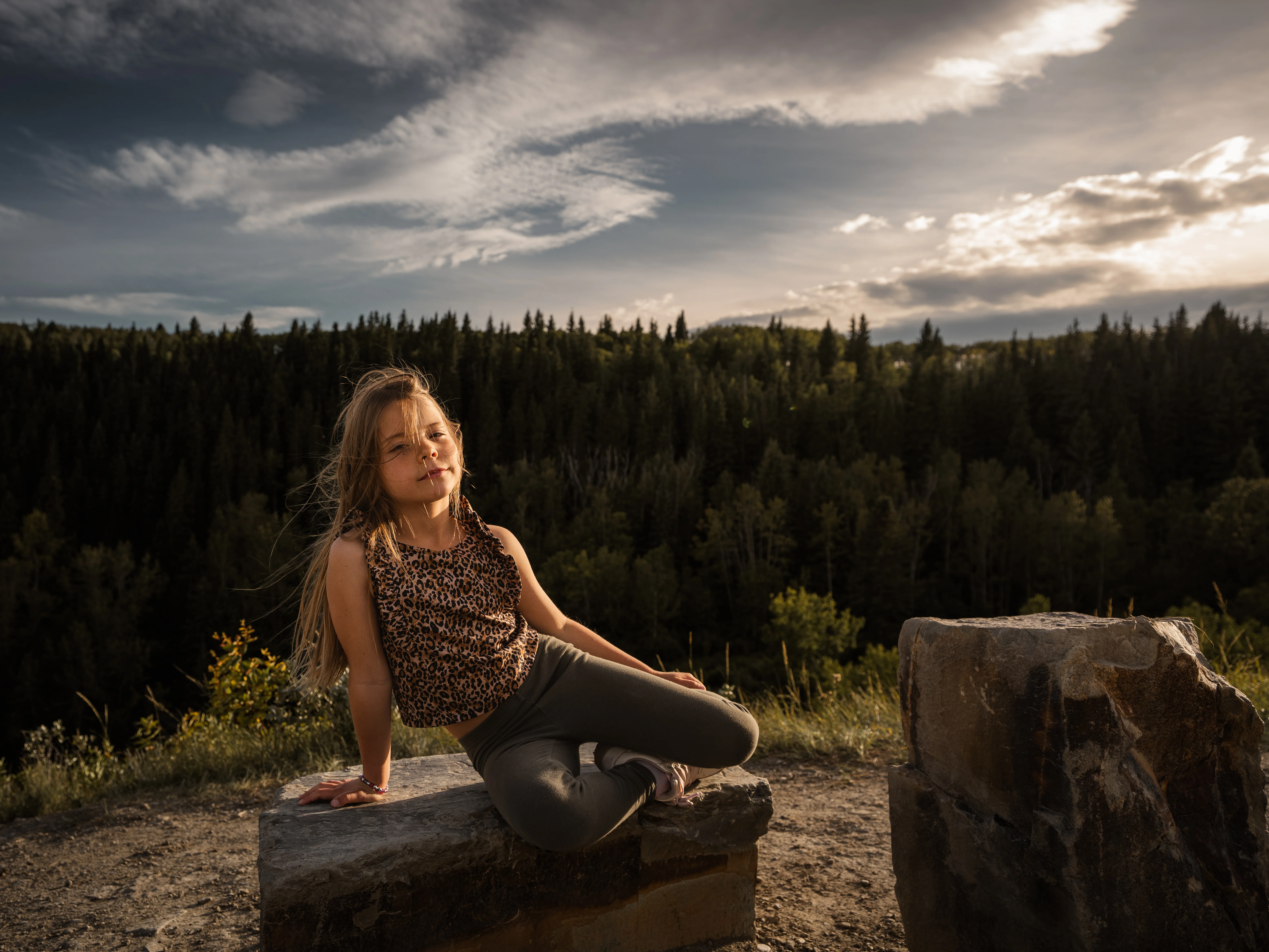 A child sitting on a rock in warm evening light, with forest and a wide Alberta sky behind her.